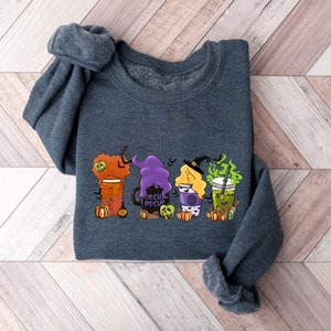 Puede incluir: Una sudadera gris oscuro con un gr&aacute;fico de tres tazas de caf&eacute; con tem&aacute;tica de Halloween. Las tazas est&aacute;n decoradas con brujas, calabazas y el texto "Hocus Pocus".