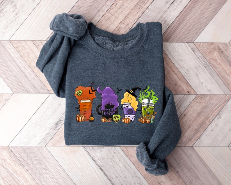 Puede incluir: Una sudadera gris oscuro con un gr&aacute;fico de tres tazas de caf&eacute; con tem&aacute;tica de Halloween. Las tazas est&aacute;n decoradas con brujas, calabazas y el texto "Hocus Pocus".