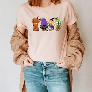 Puede incluir: Una camiseta rosa con un gr&aacute;fico de cuatro brujas vestidas como diferentes tipos de bebidas. Las brujas sostienen tazas de latte de calabaza especiada, un caldero, una poci&oacute;n y una bebida de t&eacute; verde. El texto "Boo Crew" est&aacute; en el caldero.