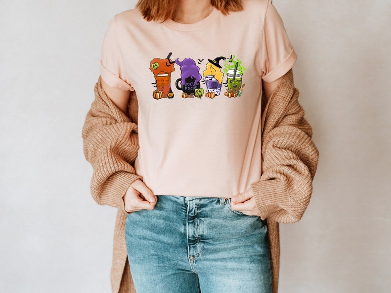 Puede incluir: Una camiseta rosa con un gr&aacute;fico de cuatro brujas vestidas como diferentes tipos de bebidas. Las brujas sostienen tazas de latte de calabaza especiada, un caldero, una poci&oacute;n y una bebida de t&eacute; verde. El texto "Boo Crew" est&aacute; en el caldero.