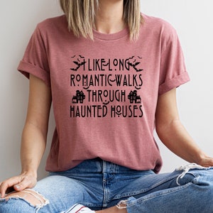 以下が含まれることがあります： 薄紫色のTシャツに、黒いグラフィックで「長いロマンチックな散歩は、幽霊屋敷の中を歩くのが好き」と書かれています。