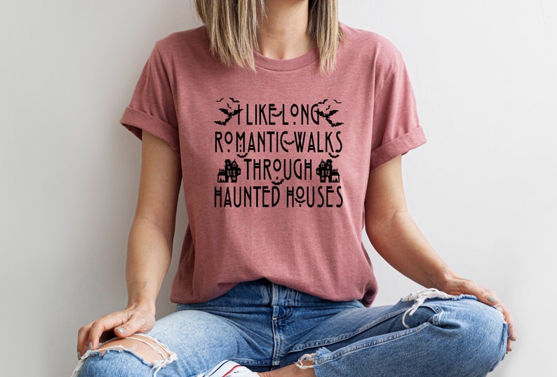 以下が含まれることがあります： 薄紫色のTシャツに、黒いグラフィックで「長いロマンチックな散歩は、幽霊屋敷の中を歩くのが好き」と書かれています。