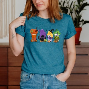 Puede incluir: Una camiseta azul turquesa con un gr&aacute;fico de cuatro vasos de caf&eacute; con tem&aacute;tica de Halloween. Los vasos est&aacute;n decorados con brujas, calabazas y el texto "Hocus Pocus".