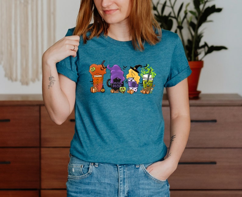 Puede incluir: Una camiseta azul turquesa con un gr&aacute;fico de cuatro vasos de caf&eacute; con tem&aacute;tica de Halloween. Los vasos est&aacute;n decorados con brujas, calabazas y el texto "Hocus Pocus".