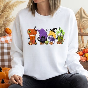 Puede incluir: Sudadera blanca con un gr&aacute;fico de cuatro bebidas con tem&aacute;tica de Halloween. Las bebidas est&aacute;n decoradas con brujas, calabazas y el texto "Hocus Pocus".
