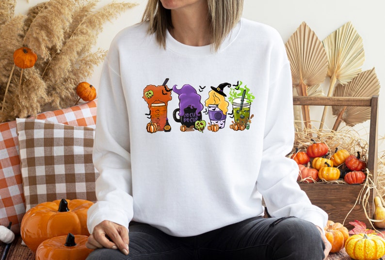 Puede incluir: Sudadera blanca con un gr&aacute;fico de cuatro bebidas con tem&aacute;tica de Halloween. Las bebidas est&aacute;n decoradas con brujas, calabazas y el texto "Hocus Pocus".