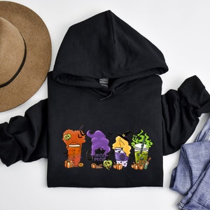 Puede incluir: Sudadera con capucha negra con un gr&aacute;fico de tres brujas sosteniendo tazas de caf&eacute; con el texto "Hocus Pocus" en una de las tazas.