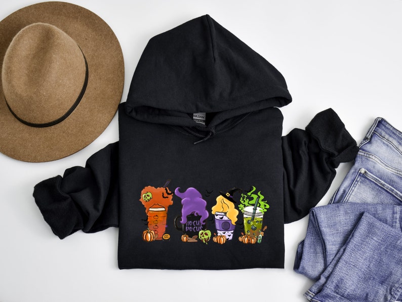 Puede incluir: Sudadera con capucha negra con un gr&aacute;fico de tres brujas sosteniendo tazas de caf&eacute; con el texto "Hocus Pocus" en una de las tazas.