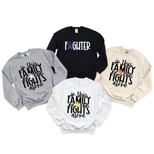 May include: Four sweatshirts with different colors and designs. The sweatshirts have the text "in this family no one fights alone" and a ribbon graphic. One sweatshirt is black with the text "fighter" and a ribbon graphic.