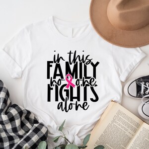 May include: White t-shirt with black text that reads "in this family no one fights alone" with a pink ribbon graphic.