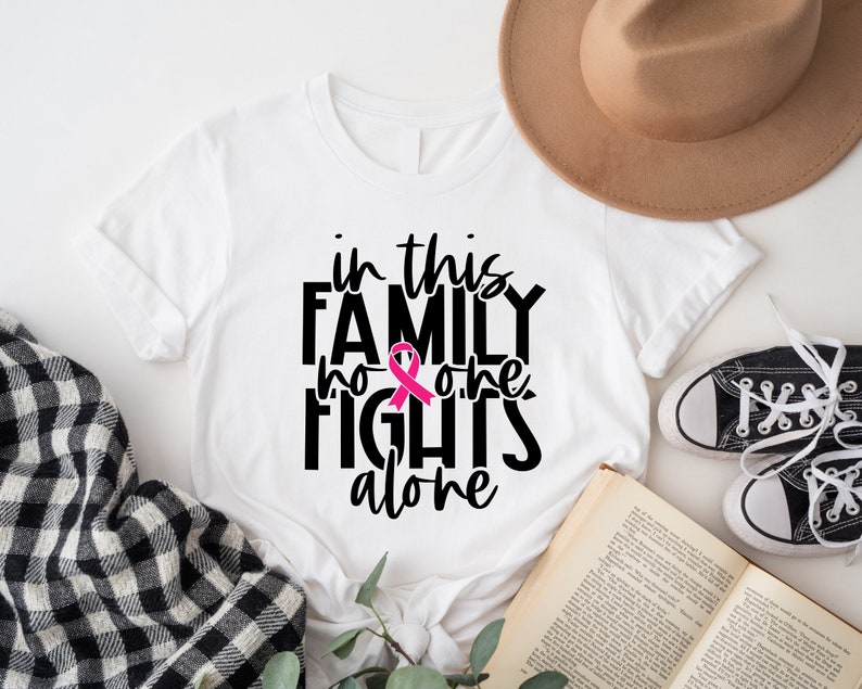 May include: White t-shirt with black text that reads "in this family no one fights alone" with a pink ribbon graphic.