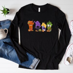 Puede incluir: Camiseta negra de manga larga con un gr&aacute;fico de cuatro tazas de caf&eacute; con tem&aacute;tica de Halloween. Las tazas est&aacute;n decoradas con sombreros de bruja, calabazas y el texto "Hocus Pocus".