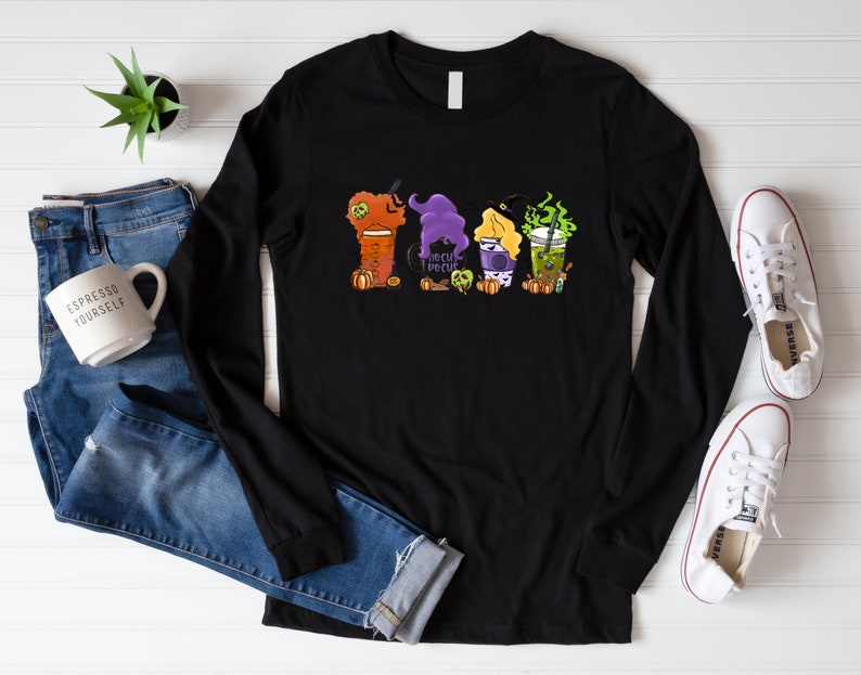 Puede incluir: Camiseta negra de manga larga con un gr&aacute;fico de cuatro tazas de caf&eacute; con tem&aacute;tica de Halloween. Las tazas est&aacute;n decoradas con sombreros de bruja, calabazas y el texto "Hocus Pocus".