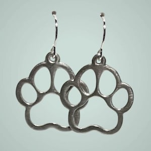 May include: A pair of silver-toned drop earrings featuring a cutout paw print design. Each earring consists of a metal paw shape suspended from a curved wire hook, showcasing a smooth, metallic finish and a hollow, open-frame construction.