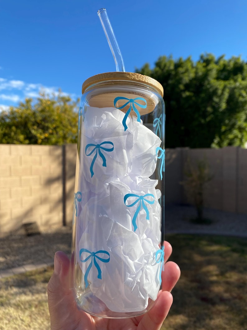 May include: A clear glass water bottle with a bamboo lid and a blue bow pattern. The bottle has a glass straw.