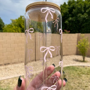 May include: A clear glass reusable water bottle with a pink bow pattern and a bamboo lid. The bottle has a glass straw.
