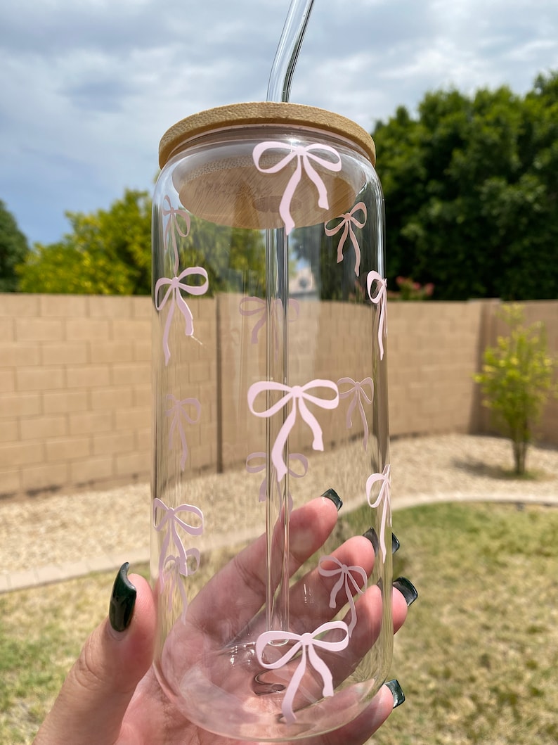 May include: A clear glass reusable water bottle with a pink bow pattern and a bamboo lid. The bottle has a glass straw.