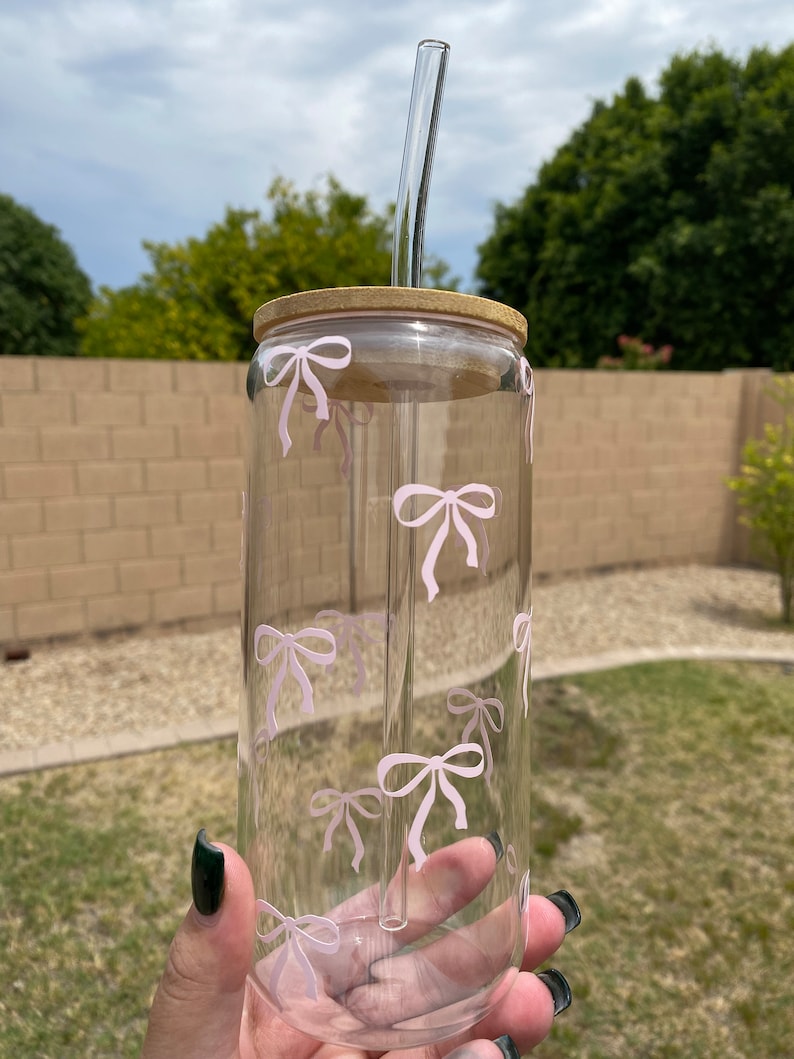 May include: A clear glass tumbler with a wooden lid and a pink bow pattern. The tumbler has a glass straw.