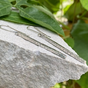 May include: A pair of silver-toned drop earrings featuring long, slender rectangular bars with a textured, weathered surface finish. Each earring is attached to a simple curved ear wire hook, creating a linear and minimalist dangling design.