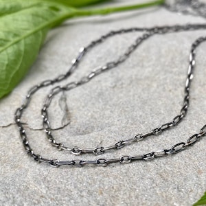 May include: A dark, oxidised silver chain necklace featuring elongated, rectangular links. The metal has a matte, gunmetal-toned finish with visible texture on the surface of each individual link, creating a continuous, flexible chain design.