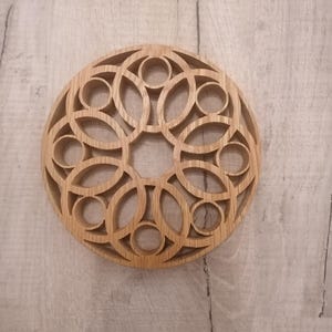 Könnte beinhalten: Ein Untersetzer aus Holz mit einem runden Design mit filigranen Ausschnitten in Form von Kreisen und Ovalen. Der Untersetzer ist aus hellbraunem Holz gefertigt und hat eine natürliche Oberfläche.