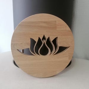 May include: A round, wooden trivet with a carved lotus flower design. The trivet is made of light-colored wood and has a natural finish. The lotus flower is cut out of the wood, creating a negative space. The trivet is designed to protect surfaces from heat.