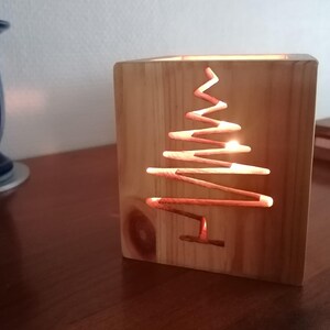 May include: A wooden candle holder with a Christmas tree design cut out. The candle holder is square and made of light-colored wood. A lit candle inside illuminates the tree shape, creating a warm glow. The candle holder is on a wooden surface.