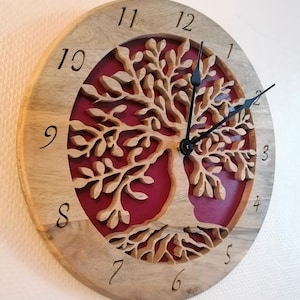 May include: A round wooden wall clock with a tree of life design. The clock face features a light brown wooden frame with carved numbers and a red background. The tree is carved from light brown wood, and the clock hands are black.