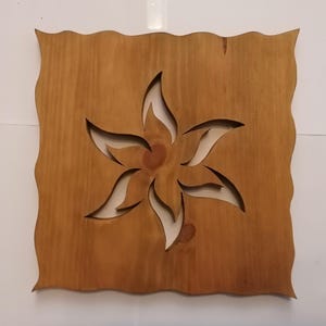 Puede incluir: Una pieza de arte mural cuadrada de madera con un diseño de flor recortado en el centro. La madera tiene un color marrón claro natural con un borde ondulado. La flor tiene seis pétalos y un centro circular.