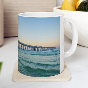 Op de afbeelding: Witte keramische mok met een kustscène van een pier die zich uitstrekt in de zee onder een blauwe en oranje lucht. De mok staat op een beige onderzetter, met een vage achtergrond van keukenartikelen.