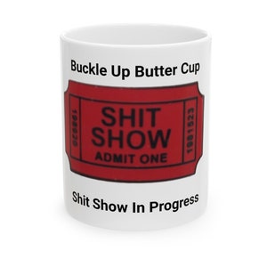 May include: White ceramic coffee mug with black text that reads "Buckle Up Butter Cup" and a red ticket with black text that reads "Shit Show Admit One". The bottom of the mug reads "Shit Show In Progress".