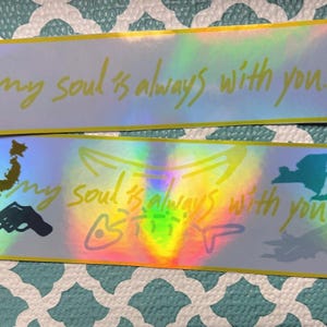 May include: Two holographic stickers with the text "my soul is always with you" in a handwritten font. One sticker has additional illustrations and text. The stickers have a yellow border and are on a blue patterned surface.