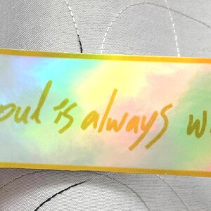 May include: A rectangular sticker with a holographic background and gold trim. The sticker features the handwritten text, "my soul is always with you." The sticker is held by a person.