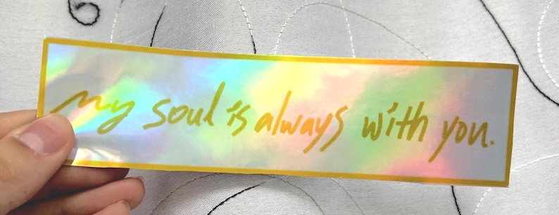 May include: A rectangular sticker with a holographic background and gold trim. The sticker features the handwritten text, "my soul is always with you." The sticker is held by a person.