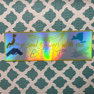 May include: Holographic sticker with a gold border. The sticker has the text "my soul is always with you" and illustrations of a gun, a map of Japan, a New York state outline, and a stylized drawing of a creature.
