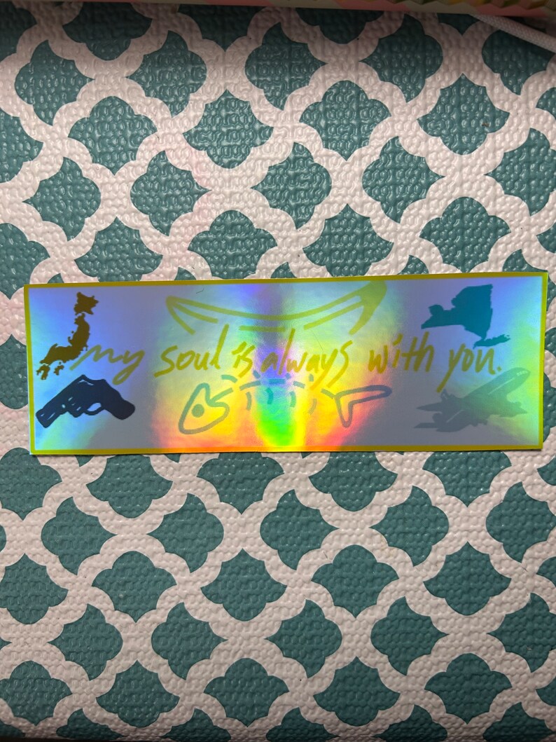 May include: Holographic sticker with a gold border. The sticker has the text "my soul is always with you" and illustrations of a gun, a map of Japan, a New York state outline, and a stylized drawing of a creature.