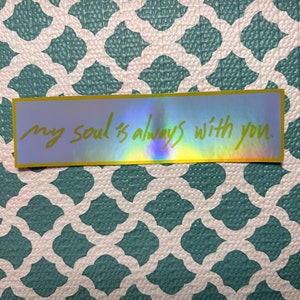 May include: A rectangular sticker with a holographic finish and a yellow border. The sticker features the handwritten text, "my soul is always with you." The sticker is placed on a teal and white patterned surface.