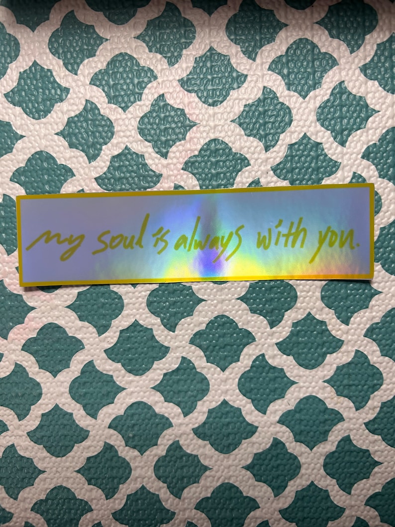 May include: A rectangular sticker with a holographic finish and a yellow border. The sticker features the handwritten text, "my soul is always with you." The sticker is placed on a teal and white patterned surface.