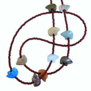 May include: A necklace featuring carved bear pendants in various colors, including green, beige, and blue, strung on a dark red beaded chain. The necklace has a unique, handcrafted appearance.