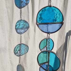 May include: Stained glass suncatcher with a variety of blue and teal glass shapes. The suncatcher is made of multiple pieces of glass, connected by metal, and hangs from a chain. The glass has a textured appearance.