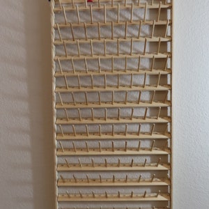 May include: A wooden thread spool rack with 72 pegs for storing thread spools. The rack is mounted on a wall and has two red plastic stoppers at the top.