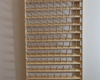 May include: A wooden thread spool rack with 72 pegs for storing thread spools. The rack is mounted on a wall and has two red plastic stoppers at the top.