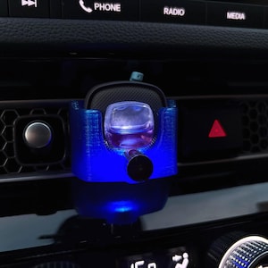 May include: A blue car air freshener holder with a clear, square-shaped container filled with a pink liquid. The holder is mounted on a car's dashboard, with a blue light illuminating the holder and the liquid.