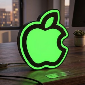 May include: A bright neon-style sign shaped like an apple logo with a vibrant green illuminated centre and a thick black border. The sign features a black power cord with an inline switch and rests on a surface with a small metallic plaque.