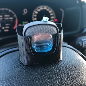 May include: A black 3D-printed car accessory holder with a gray cushioned top and a clear, blue-tinted, square-shaped object. The holder is mounted on a black leather steering wheel, with the car's dashboard visible in the background.