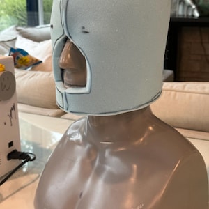 Puede incluir: Un cubrecabezas de espuma azul claro con un recorte central para la nariz y la boca, que presenta costuras oscuras visibles a lo largo de la parte superior y los lados. La máscara de espuma se muestra sobre un busto de maniquí gris liso.