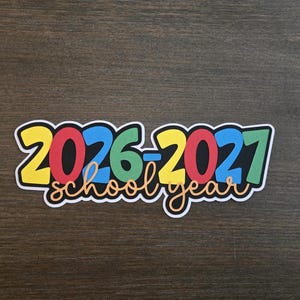 May include: A die-cut sticker featuring the text 2026-2027 in bold, multi-colored numbers including yellow, red, blue, and green, with the words school year written in a cursive, orange font underneath, all outlined in a thick white border.