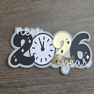 May include: A layered papercraft design for New Year 2026. The design features the numbers "2026" with a clock face and the words "new year". The numbers are black and gold, with diamond accents. The clock hands point to midnight.