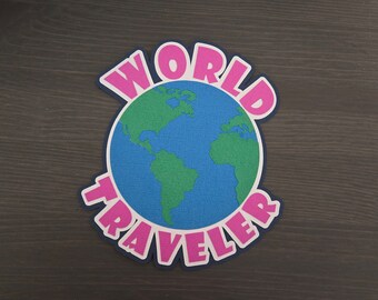 May include: A colorful, layered paper cutout featuring a blue globe with green continents. The words "WORLD TRAVELER" are in pink, outlined in white, and set against a dark blue background. The design is likely for travel-themed products.