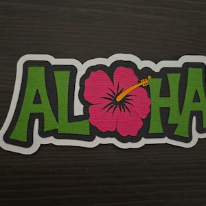 Puede incluir: Un letrero de "Aloha" verde y negro con una flor de hibisco rosa en el centro. El letrero tiene un borde blanco.